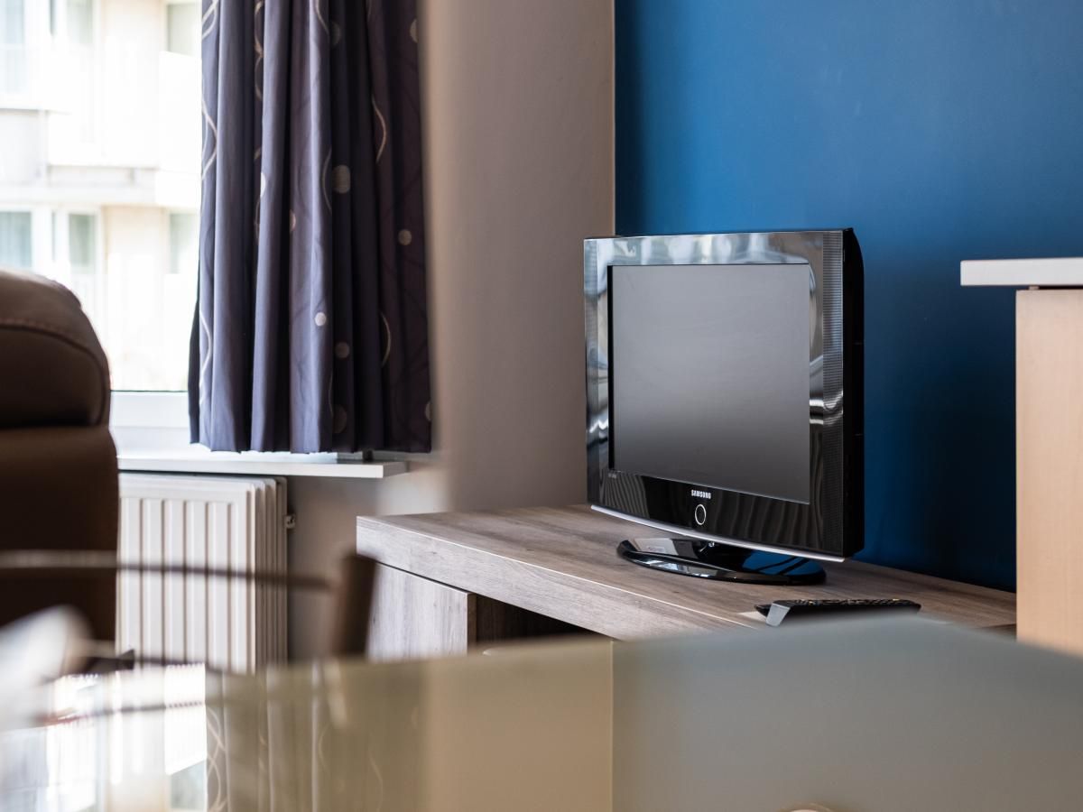 Gezellige woonkamer in Apollo Gelijkvloers vakantiehuis, Koksijde, Belgie, met moderne tv op houten meubel en blauwe muur.