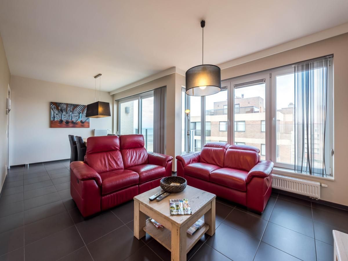 Gezellige woonkamer in De Dijk zesde vakantiehuis, Koksijde, met comfortabele rode banken en veel natuurlijk licht.
