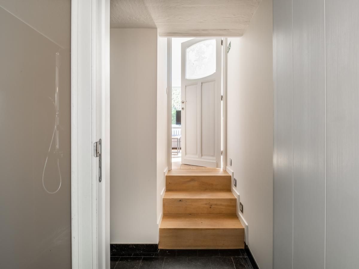 Lichte hal van vakantiehuis Tranquil in Koksijde, West-Vlaanderen, met houten trap en open deur naar een zonnig terras.