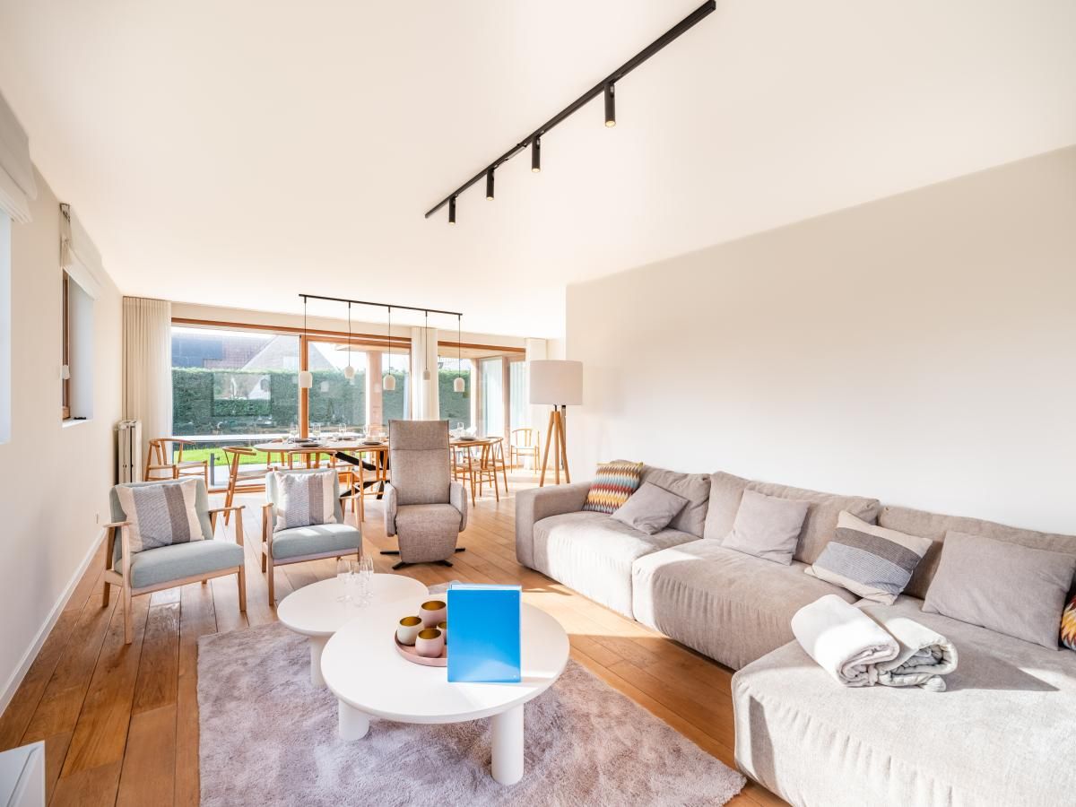 Helles Wohnzimmer des Ferienhauses "Pure Relax" in Oostduinkerke, Belgien, mit gemuetlicher Sitzecke und Blick auf den Garten.
