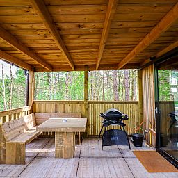 Overdekte veranda van Comfort Classic in Neeroeteren, Limburg, Belgie, met houten zitgedeelte en barbecue, omgeven door natuur.